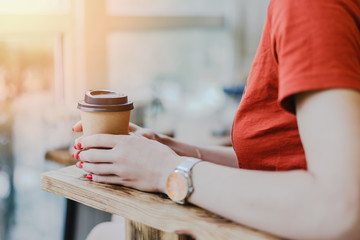 Paper coffee cup to go in woman's hands with red manicure while sitting in cafe.