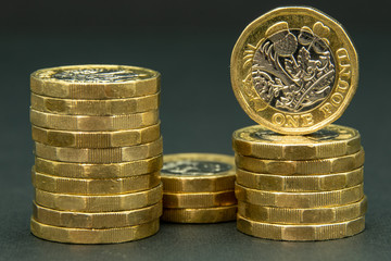 Three stacks of British one pound coins with one coin standing facing front on one of the stacks - all on a black background