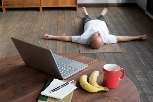 Blurred phot of senior caucasian man meditating on a wooden floor of living room and lying in Shavasana pose after practice. Table with laptop, banana and cup on the foreground