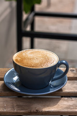 Blue round cup of latte coffee on a plate on the cafe wooden table outside.