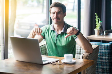 Dislike! Young dissatisfied businessman in green t-shirt sitting, working on laptop, looking at camera and showing thumbs down. business and freelancing concept. indoor shot near big window at daytime