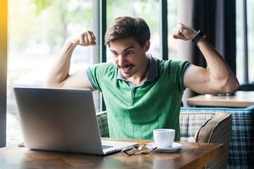 I am strong! Young aggressive businessman in green t-shirt sitting, looking at laptop screen on video call, showing his biceps muscle, angry face. business concept. indoor shot near window at daytime.