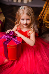 little blonde girl in evening red dress near the Christmas tree, child with Christmas gifts, the night before Christmas