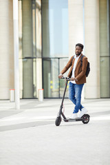 Full length portrait of trendy African man riding electric scooter and looking at camera while commuting in city street, copy space