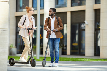 Full length portrait of contemporary young people riding electric scooters in city street and smiling happily looking at each other, copy space