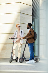 Full length portrait of modern young couple riding electric scooters in city street and chatting, copy space