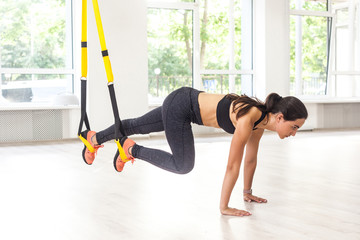 Side view portrait of young muscular woman wearing black sportswear standing on plank position use fitness straps, doing push ups while legs hanging on trx. Indoor, window background, abs exercises