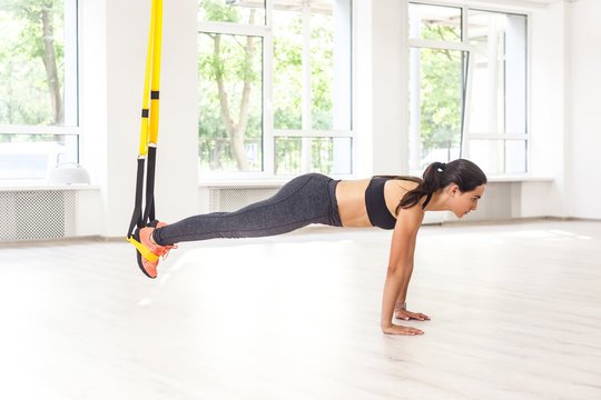 Side view portrait of young muscular woman wearing black top and leggings standing on plank position use fitness straps, doing push-ups while legs hanging on trx. Indoor, window background