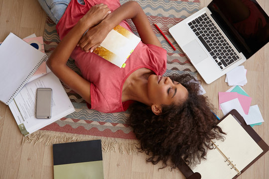 Beautiful young curly lady with dark skin lying on floor between books, notebooks and laptop, posing over colored carpet with closed eyes and pleasant smile
