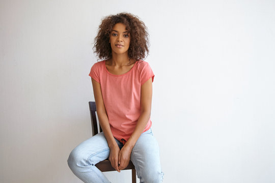 Indoor photo of attractive curly woman with dark skin posing over white wall in casual clothes, looking at camera with calm face and smiling slightly