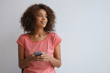 Inoor portrait of beautiful curly woman with dark skin posing over white background, looking aside with pleasant smile, holding smartphone in hands