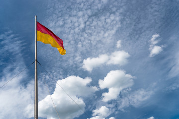 Flag in red at the top and yellow at the bottom, hanging on the mast, waving in the wind. In the background a blue sky with white clouds.
