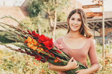 Outdoor portrait of young beautiful woman in summer garden, holding bouquet of gladiolus flowers - obrazy, fototapety, plakaty