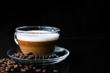 Transparent glass cup hot cappuccino with visible layers of coffee, milk and foam and coffee beans on black wood table at cafe. Dark scene. Place for text. Stylish toning, smoke