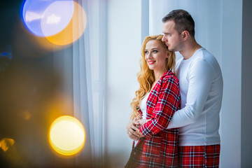 Loving happy couple husband and wife in plaid pajamas are stand near a huge window and hugging. Plastic windows, a warm comfortable house.