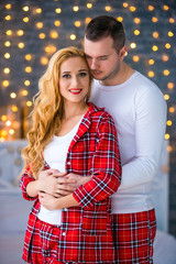 Loving happy couple husband and wife in plaid pajamas stand near the christmas lights of garlands. Festive mood. Merry christmas and happy new year.