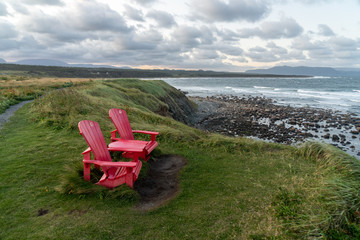 Fototapeta premium A pair of adirondack chairs at the edge of a cliff overlooking the ocean, on a patch of green grass and a hiking trail nearby