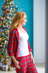 Beautiful blonde woman in red checkered pajamas stands near the window and demonstrates comfortable home clothes.