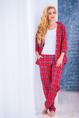 Beautiful blonde woman in red checkered pajamas stands near the window and demonstrates comfortable home clothes.