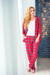 Beautiful blonde woman in red checkered pajamas stands near the window and demonstrates comfortable home clothes.