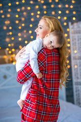 A beautiful young mother in checkered pajamas holds a little son in her arms. Caring and motherly love.