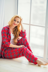 Beautiful blonde woman in red checkered pajamas sits near the window and demonstrates comfortable home clothes.