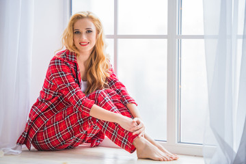 Beautiful blonde woman in red checkered pajamas sits near the window and demonstrates comfortable home clothes.