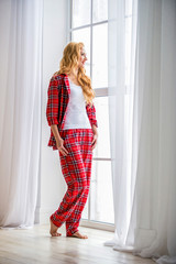 Beautiful blonde woman in red checkered pajamas stands near the window and demonstrates comfortable home clothes.