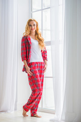 Beautiful blonde woman in red checkered pajamas stands near the window and demonstrates comfortable home clothes.