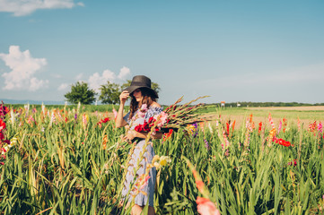 Gorgeous young woman picking flowers in a field, wearing summer dress, black straw hat