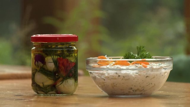 Steady, medium close up shot of a jar containing olive oil, spices, garlic and a flower. A bowl of dip sits beside.
