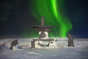 A traditional stone inukshuk, an Inuit cultural symbol used as a landmark to guide travelers in the far north, showing they are on the right path. Shimmering green aurora borealis in the background.