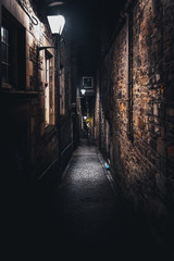 A dark creepy narrow European alley at night, surrounded by bricks and cobblestone. Illuminated only with some street lamps. Concept of scared or being alone and frightened