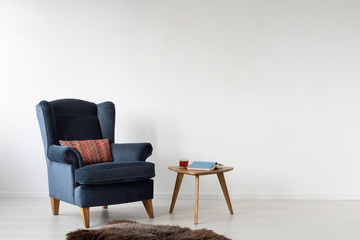 Navy blue armchair next to wooden table in white flat interior with copy space on the wall. Real photo
