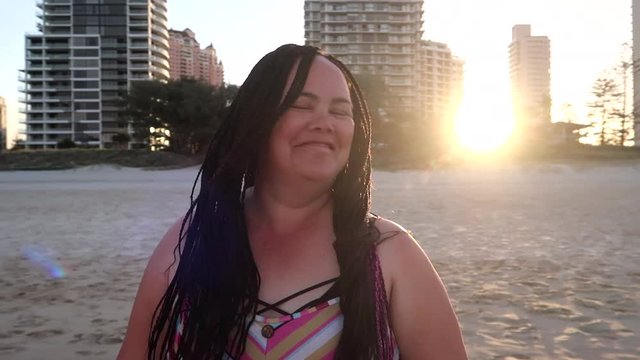 A Maori woman on the Gold Coast of Australia plays with her boxed braided hair. The setting sun behind her casts lens flares across the image as the camera moves around her.