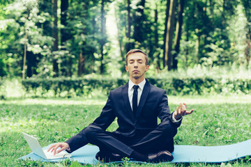 Obraz premium Man in a suit with tie. Business man relaxing in a park in the lotus position, he can't relax in any way, typing text on a computer with one hand. one eye mows into the monitor.