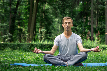 Young attractive man in sport clothes is meditating in the lotus position with a pacified face in the park against the background of green grass and trees.