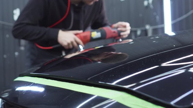 A male polisher works on polishing a black car in a well-lit but shabby garage