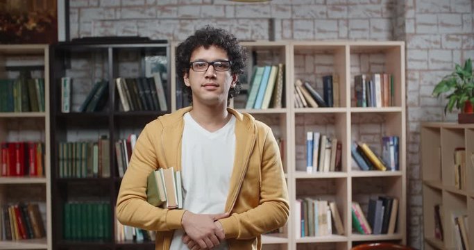Asian guy with curly hair is standing between bookshelves of library holding books in hands, looking at camera and smoling - portrait closeup 4k, education, student lifestyle concept 4k