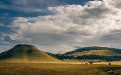 Grass hills covered with trees in steppe under spectacular clouds sky during sunset at Khakassia, Siberia, Russia.