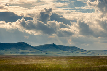 Grass hills on horizon in steppe under heavy clouds sky during sunset with sun light beams at Khakassia, Siberia, Russia.