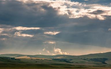Grass hills on horizon in steppe under heavy clouds sky during sunset with sun light beams at Khakassia, Siberia, Russia.