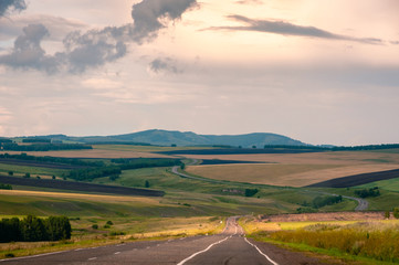 Sunset landscape with agricultural fields in steppe with forest and curvy asphalt road to the horizon at Khakassia, Siberia, Russia.