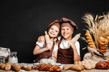 Adorable girl with brother in chief hats and aprons cooking at table with bread loaves making fresh dough and having fun