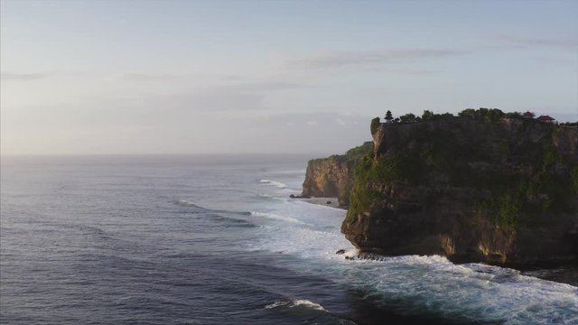 Aerial view of amazing high cliffs above blue ocean. Foamy waves. Twilight. Silhouette of Uluwatu temple on the cliff top. Bali, Indonesia