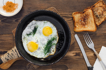 Pan of fried eggs with toasts and fork on dark brown wooden table, top view