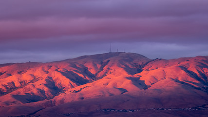 Sunset landscape in South San Francisco bay with storm clouds covering the sky; mountains slopes and peaks (part of the Diablo mountain range) colored red by the setting sun; San Jose, California