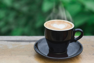 Hot coffee with smoke, heart shape with milk latte on top of black ceramic cup on wooden table in coffee shop with green background, copy space for your text. Times to relax with caffiene concept.