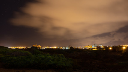 The skyline of La Mata, part of the Spanish city of Torrevieja, in the early morning in the dark. The small pine forest is right on the beach. The city and the clouds in the sky are brightly lit.