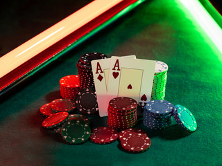 Two aces standing leaning on chips piles, some of them laying nearby on green cover of playing table, under green and red neon lights. Close-up.